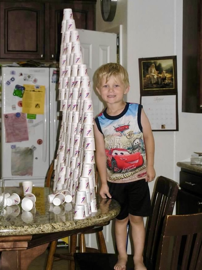 Young boy showing off the paper cup structure he built. The structure is taller than he is.